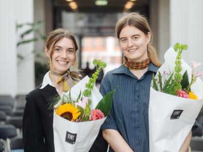 De to smilende prisvindere Helene Franch og Feline Kamber holder blomsterbuketter foran rækker af stole.